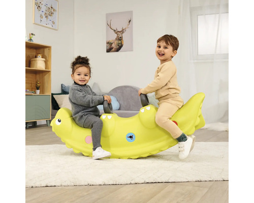 Zwei Kinder sitzen auf einer grünen Krokodilwippe in einem hellen Raum.