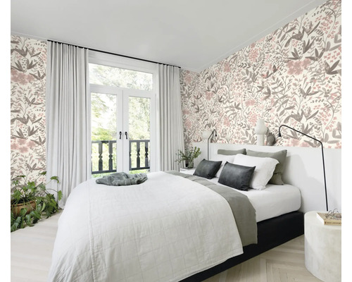 Schlafzimmer mit Bett vor einer floral gemusterten Tapete und einem Fenster mit Vorhängen.