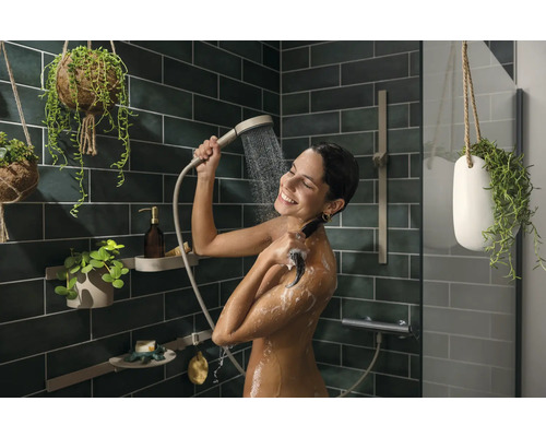 Frau benutzt Handbrause in einer Dusche mit grünen Fliesen und hängenden Pflanzen