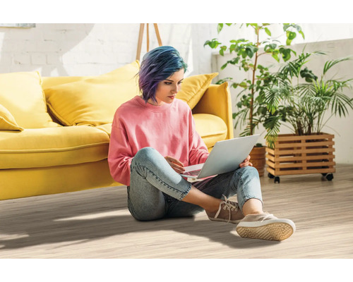 Frau sitzt auf dem Boden und benutzt einen Laptop, im Hintergrund Sofa und Pflanzen.