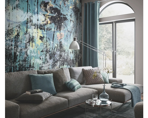 Wohnzimmer mit grauer Couch, Kissen, Tischlampe, Vorhang und einer Wandtapete mit abstraktem Design