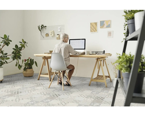 Eine Frau sitzt an einem Schreibtisch mit einem Computer in einem hellen Raum mit Pflanzen und gemustertem Bodenbelag.