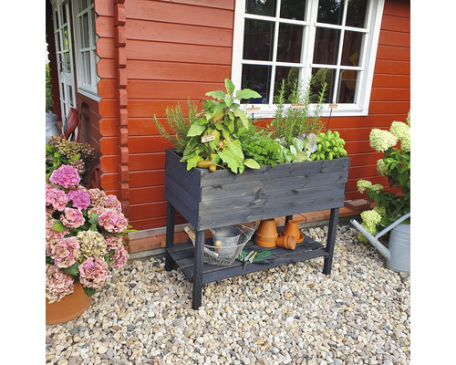Hochbeet aus Holz mit Kräutern, Ablagefläche und Gartengeräten vor einem roten Holzhaus.
