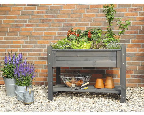 Hochbeet mit Pflanzen, Kräutern und Gartenutensilien vor einer Ziegelwand