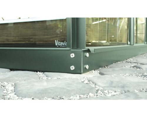 Detailansicht eines Gewächshauses mit Aluminiumrahmen und Glasscheiben auf einem Steinboden. Vitavia Logo.