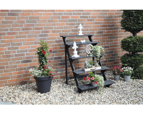 Dekorativer Gartenbereich mit Pflanzentreppe, Blumen und Zierobjekten vor einer Backsteinmauer.