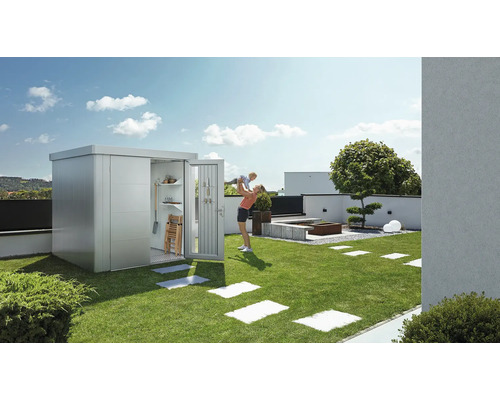 Gartenschrank aus Metall mit geöffneter Tür auf einer Rasenfläche, eine Frau hält ein Kind hoch.