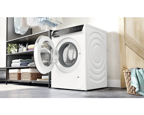 Waschmaschine mit geöffneter Tür in einem hellen Waschraum neben einem Regal mit Wäsche und Körben