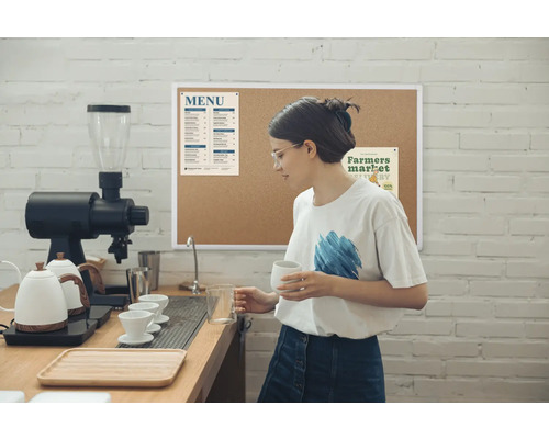 In einer hellen Café-Szene steht eine Frau mit Brille hinter der Theke, hält eine Tasse und einen Messbecher, umgeben von Kaffeemaschine, Wasserkochern und einer Menütafel an einer Ziegelwand.