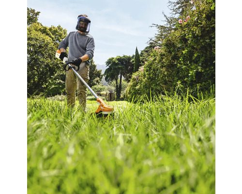 Mann mit Schutzhelm und Motorsense beim Rasenmähen im Garten.