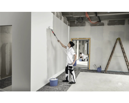 Eine Person streicht eine Wand in einem Raum mit einer Farbrolle an einem Teleskopstiel. Eine weitere Person und eine Leiter sind im Hintergrund zu sehen.