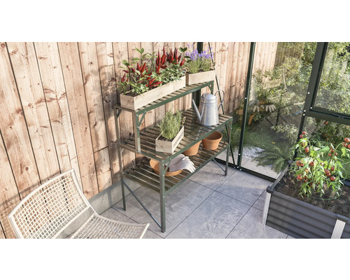 Gartenszene mit einem Pflanztisch aus Metall, bepflanzten Holzkästen, einer Gießkanne und Gartengeräten in einem Gewächshaus