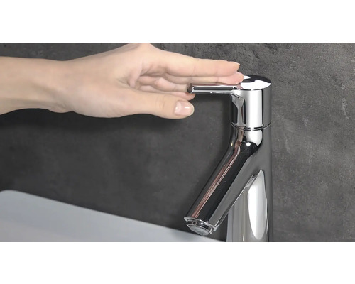 Nahaufnahme eines glänzenden, verchromten Waschtischmischers mit Bedienhebel und einer Hand, die ihn betätigt.