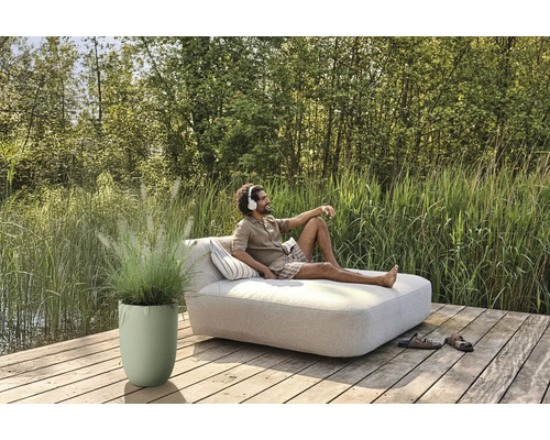 Mann entspannt mit Kopfhörern auf einer Gartenliege auf einer Holzterrasse neben einem Pflanzgefäß mit Ziergras.