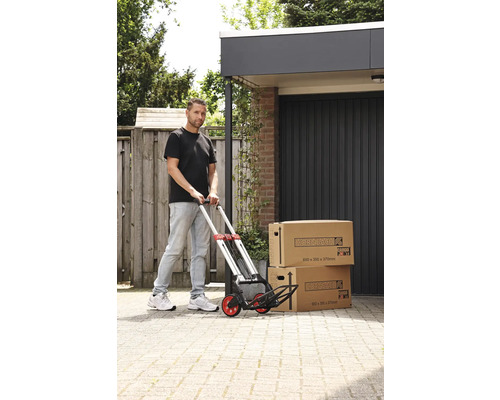 Mann schiebt einen Handwagen mit zwei Umzugskartons vor einer Garage.