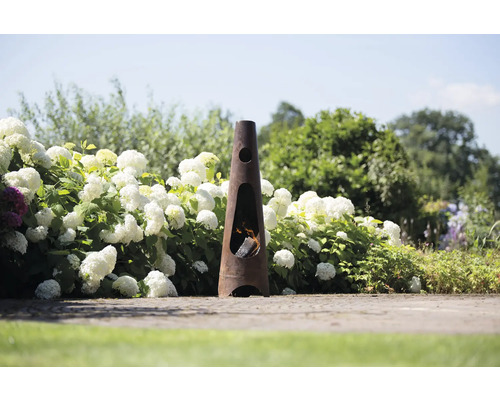 Gartenszene mit einem schlanken, kegelförmigen Kaminofen aus Metall, der mit Holz befeuert wird und von Hortensien umgeben ist.