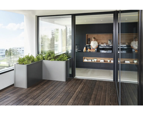 Szene mit Kräutern in grauen Pflanzkästen auf einem Holzbalkon vor einer offenen Schiebetür zu einer Küche