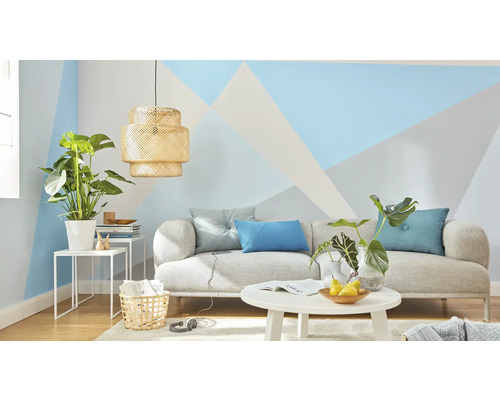 Wohnzimmer mit Sofa, Tisch, Lampe und Zimmerpflanzen vor einer Wand mit geometrischen Mustern