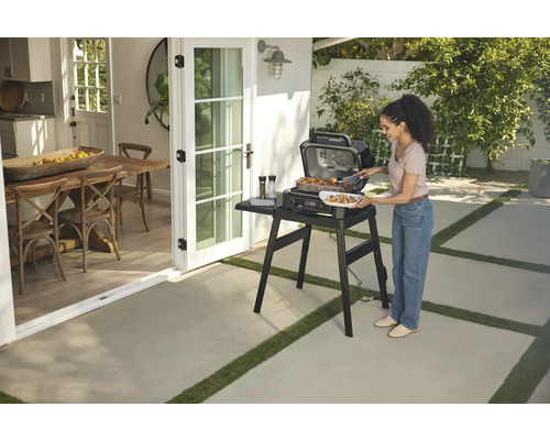 Frau grillt mit einem elektrischen Ninja Grill im Freien auf einer Terrasse neben einer offenen Tür zum Essbereich.