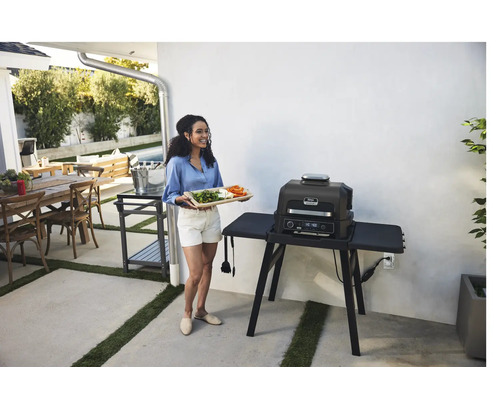 Frau mit Serviertablett neben einem elektrischen Ninja Grill im Außenbereich.