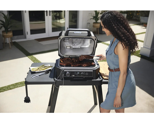 Szene mit einer Frau, die Steaks auf einem elektrischen Grill im Freien grillt, neben einem Teller Spargel.