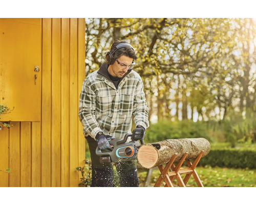 Mann sägt mit einer Akku-Kettensäge ein Holzstück auf einem Sägebock, während er eine Schutzbrille und Kopfhörer trägt.