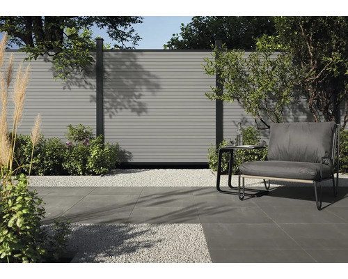 Gartenszene mit grauer Terrassendiele, Kies und Sitzgelegenheit vor einem Zaun.