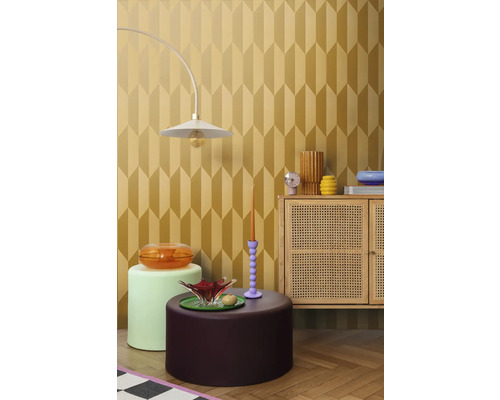 Einrichtungsbeispiel mit einer Lampe, verschiedenen Tischen und einem Schrank vor einer Tapete mit geometrischem Muster.