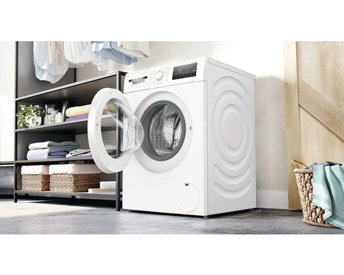 Weisse Waschmaschine mit geöffneter Tür in einem Waschraum mit Regalen und Wäschekorb.