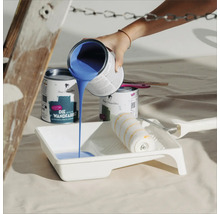 Blaue Wandfarbe wird in eine Farbwanne gegossen, daneben stehen Farbdosen und eine Farbwalze.