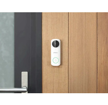 Weiße Imou Video-Türsprechanlage mit Kamera und Klingeltaste an einer hellen Holzfassade neben einer modernen dunklen Tür mit Metallgriff.
