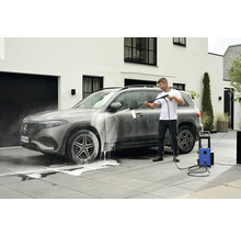 Mann reinigt ein Auto mit einem Hochdruckreiniger auf einer gepflasterten Fläche vor einem Haus.