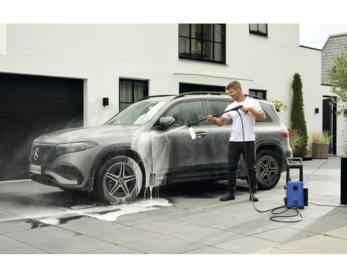 Mann reinigt ein Auto mit einem Hochdruckreiniger auf einer gepflasterten Fläche vor einem Haus.