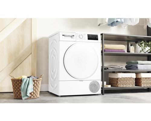 Freistehender weißer Bosch Wärmepumpentrockner in einem hellen Raum mit Wäschekorb und Regal