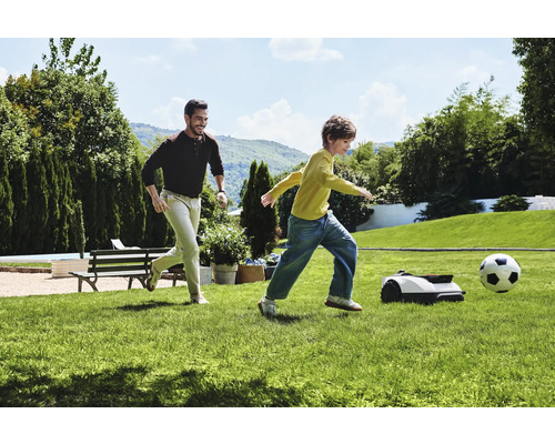 Mann und Kind spielen Fußball mit Rasenmähroboter im Garten.