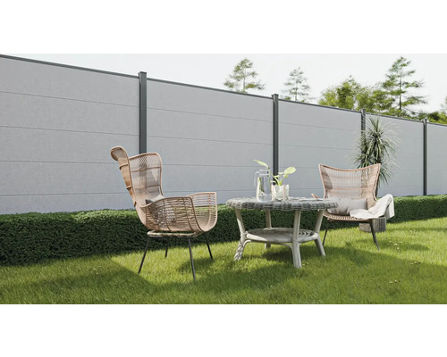 Gartenszene mit Betonzaun, Rattansesseln und rundem Tisch auf einer Rasenfläche.