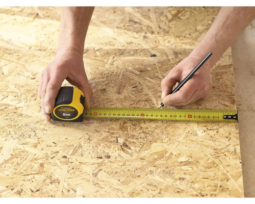 Eine Person misst ein Stück Holz mit einem Stanley Fatmax Rollbandmass von 5 Metern und markiert es mit einem Stift.