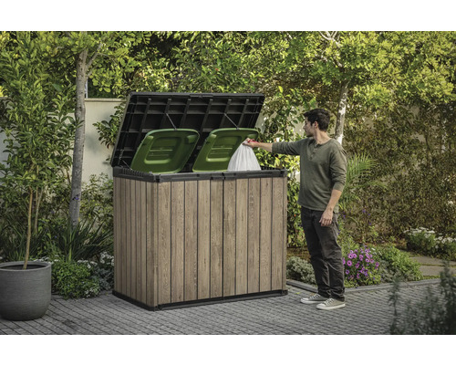 Mülltonnenbox aus Holz mit zwei grünen Mülltonnen und einer Person, die einen Müllsack hineinwirft