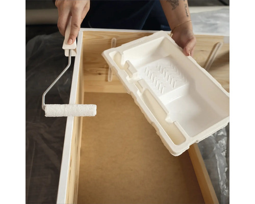 Eine Person streicht ein Holzmöbelstück mit einem Farbroller und einer Farbwanne.