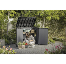 Garten mit einer Frau, die vor einem geöffneten Geräteschrank kniet, in dem Gartengeräte aufbewahrt werden.