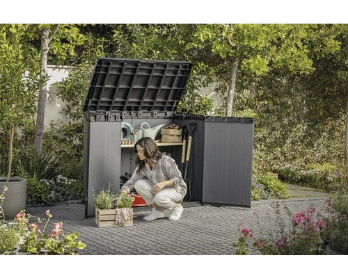 Garten mit einer Frau, die vor einem geöffneten Geräteschrank kniet, in dem Gartengeräte aufbewahrt werden.