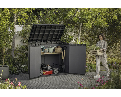 Garten Szene mit einer Frau die eine Kiste mit Pflanzen neben einem Geräteschrank trägt, der mit Gartengeräten und einem Rasenmäher gefüllt ist