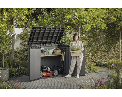 Gartenszene mit Gerätehaus, Gartengeräten und einer Frau, die eine Kiste mit Pflanzen hält.