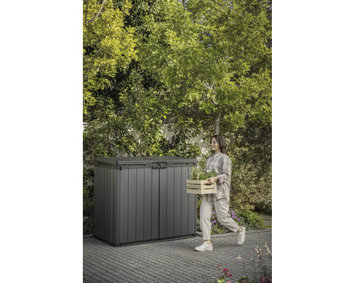 Gartenszene mit Gerätebox und Frau, die einen Holzkasten mit Pflanzen trägt