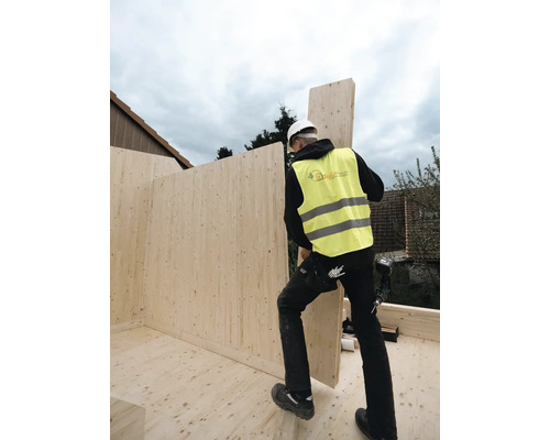 Ein Bauarbeiter mit Schutzweste und Helm montiert eine Holzwand auf einer Baustelle.
