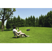Ein Mann sitzt im Garten auf einem Stuhl und bedient einen Rasenmähroboter mit seinem Smartphone.