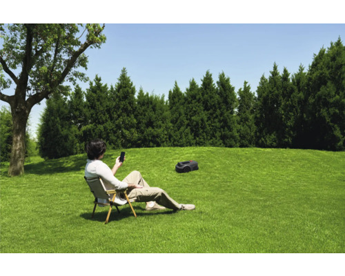 Ein Mann sitzt im Garten auf einem Stuhl und bedient einen Rasenmähroboter mit seinem Smartphone.
