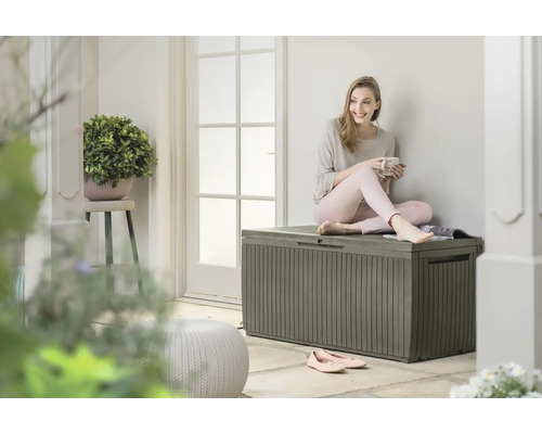 Eine junge Frau sitzt auf einer grauen Aufbewahrungsbox im Freien und hält eine Tasse