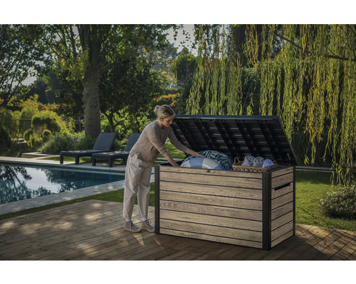 Aufbewahrungsbox Keter Ashwood 147 x 83 x 86 cm grau Eine Frau öffnet eine Gartentruhe auf einer Holzterrasse neben einem Pool und Garten.