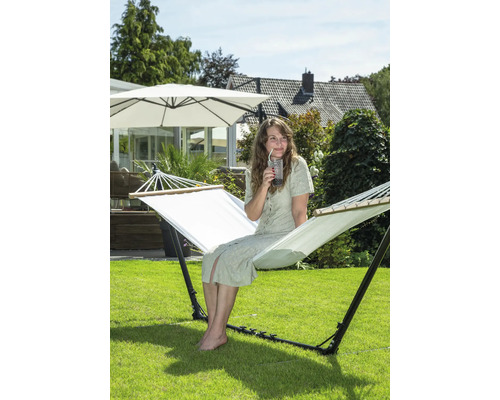 Frau sitzt in einer freistehenden Hängematte im Garten und trinkt ein Getränk.